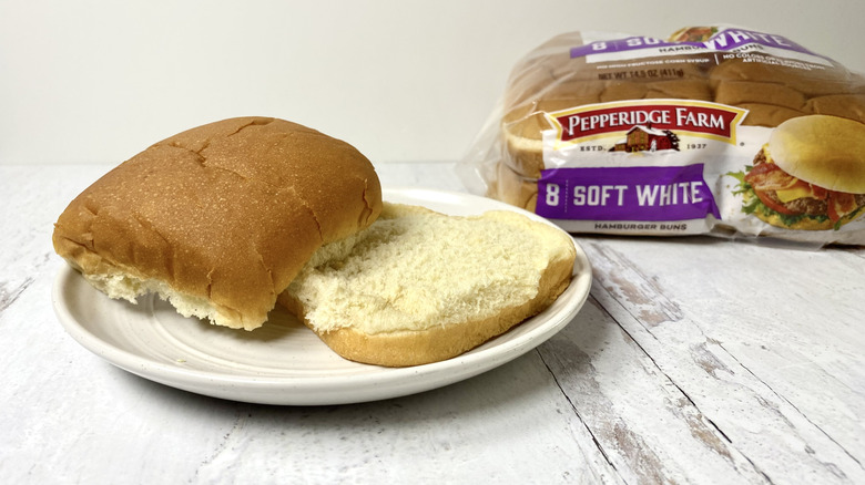 A Pepperidge Farm Soft White Hamburger Bun on a plate in front of the packaging on a white surface.