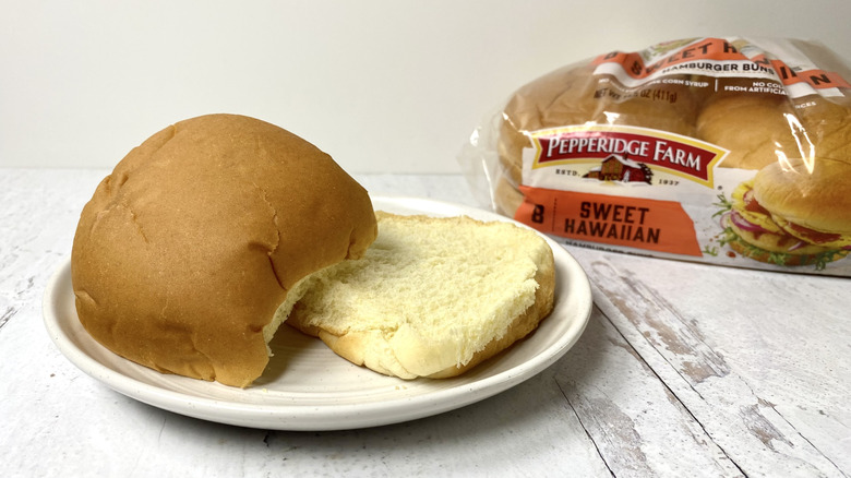 A Pepperidge Farm Sweet Hawaiian Hamburger bun on a plate in front of the packaging on a white surface.