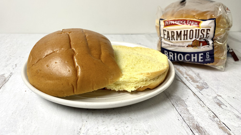 A Pepperidge Farm Farmhouse Brioche bun on a plate in front of the packaging on a white surface.