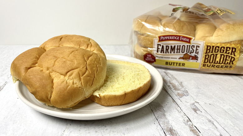 A Pepperidge Farm Farmhouse Butter Hamburger Bun on a white plate in front of its packaging on a white surface.