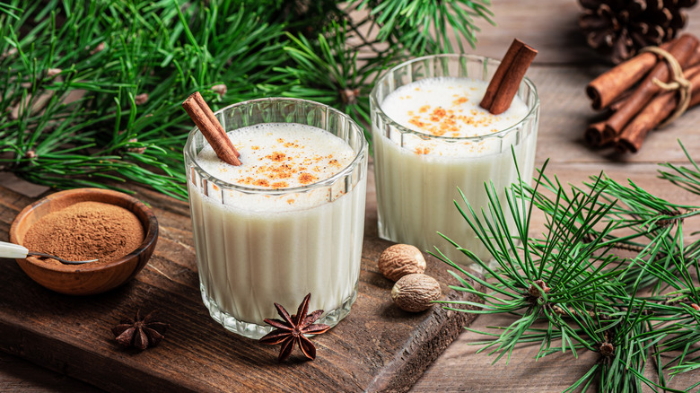 Two glasses of eggnog with cinnamon sticks in them surrounded by Christmas tree branches