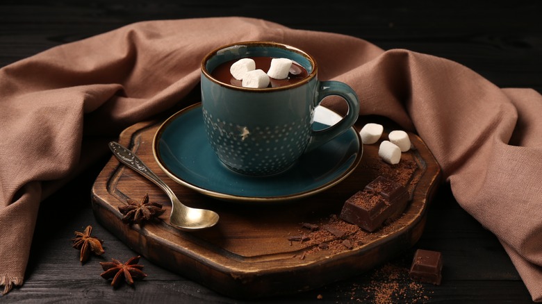 A cup of dark hot chocolate sitting on a wooden board surrounded by chocolate, marshmallows, and spices.