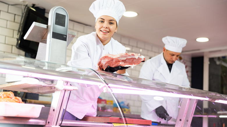 A female butcher holding up a cut of meat while a male butcher works in the background