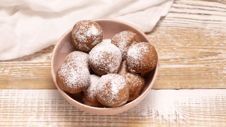 Bowl of frittelle donuts dusted in powdered sugar on wood table.