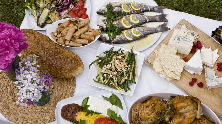 Spread of various prepared dishes and appetizers on white tablecloth with grass in background.
