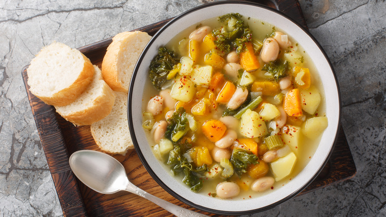 Traditional Italian vegetable and bean soup on wood tray next to spoon and bread on stone countertop.