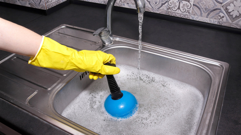 Person wearing yellow glove using plunger to unblock kitchen sink.
