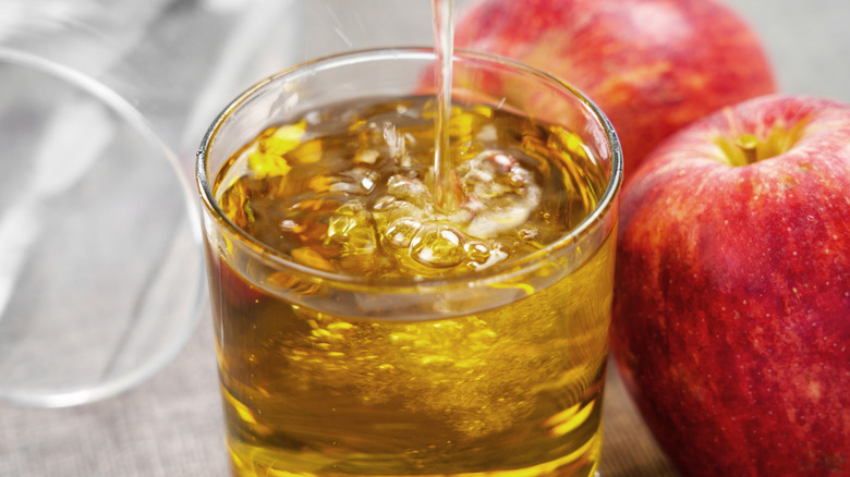 Apple cider being poured into a clear glass, with two fresh apples in the background.