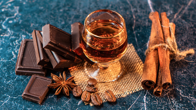 A glass of whiskey sitting next to chocolate, cinnamon sticks, coffee beans, and star anise.