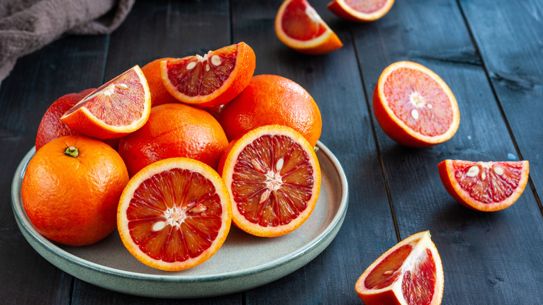 A plate containing whole blood oranges and cut blood oranges with more cut pieces surrounding the plate