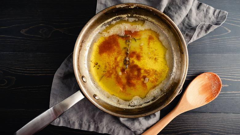 Browned butter in pan with rosemary on dish towel next to wooden spoon.