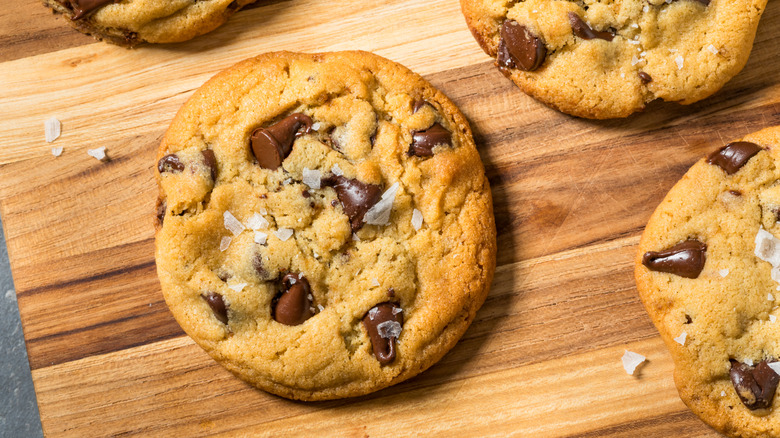Homemade chocolate chip cookies with flaky sea salt on wood cutting board.