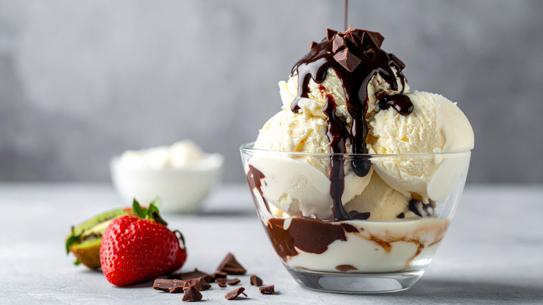 Vanilla ice cream in a glass bowl with strawberries, chocolate sauce and chopped chocolate on counter.