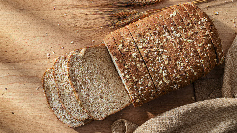 Whole grain sliced bread on wood cutting board next to dried wheat.