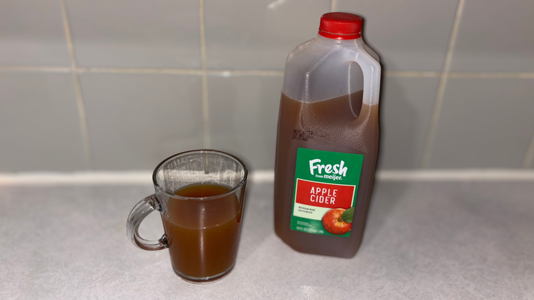 A clear mug 3/4 full of Meijer Fresh apple cider next to jug of cider.