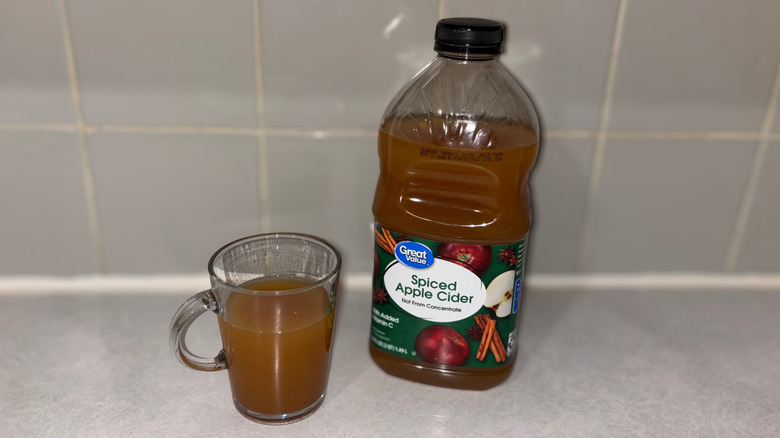 A clear mug 3/4 full of Great Value apple cider next to bottle of cider.
