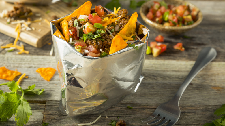 a walking taco alongside a fork on a wooden table with salsa and a cutting board in the background