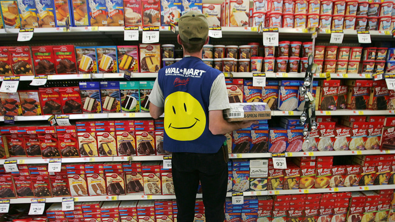 Walmart employee wearing vest with yellow smiley face faces away from camera as he restocks cake frosting