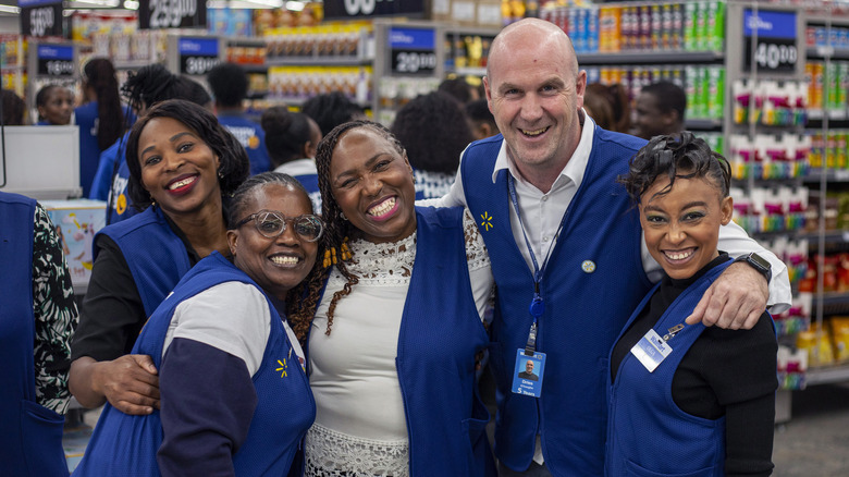 A diverse group of 5 Walmart employees, 4 women and 1 man, smile for the camera with their arms around each other