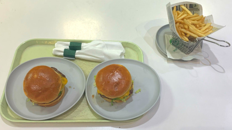 Two burgers on white plates on a yellowed tray with rolled utensils on a white table next to a small basket of fries