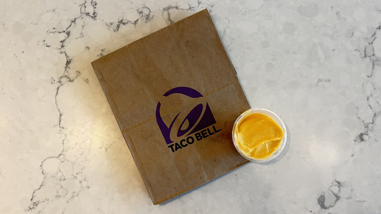 A cup of Taco Bell Nacho Cheese Sauce next to a Taco Bell bag