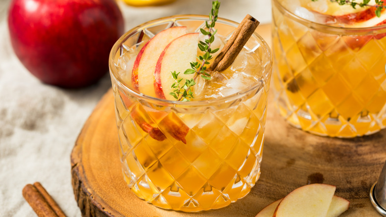 Two glasses og bourbon with apples, cinnamon sticks, and herbs.