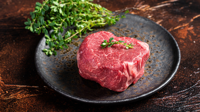 A raw filet mignon steak on a plate with herbs.