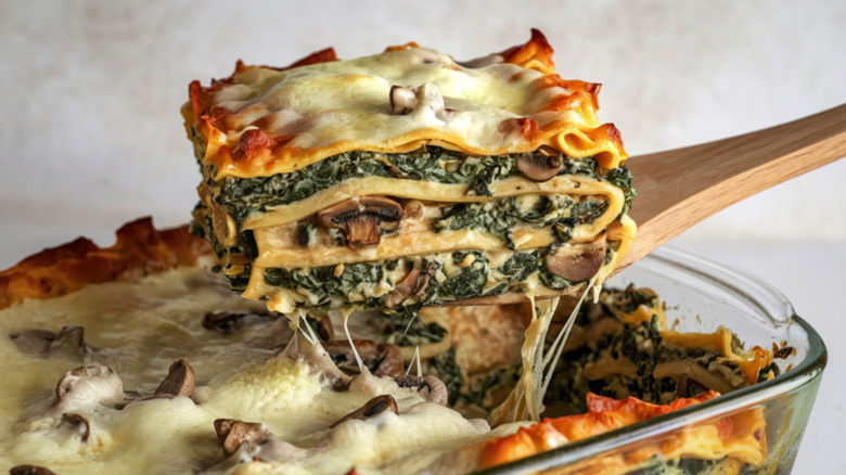 Thick spinach and mushroom lasagna lifted from a baking dish with a wooden spatula.
