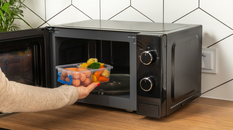 A person putting a plastic tub of sausages and vegetables into the microwave.
