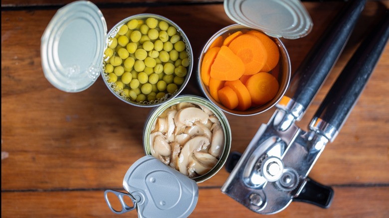 Three opened cans of vegetables next to a can opener on a wooden background