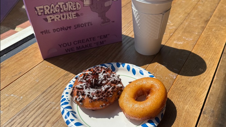 Two fresh donuts on a paper plate next to a cup of coffee on a wooden counter