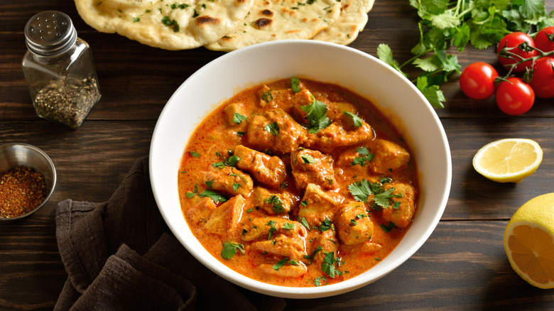 Bowl of chicken tikka masala with naan, spices, tomatoes, cilantro, and lemon on the side