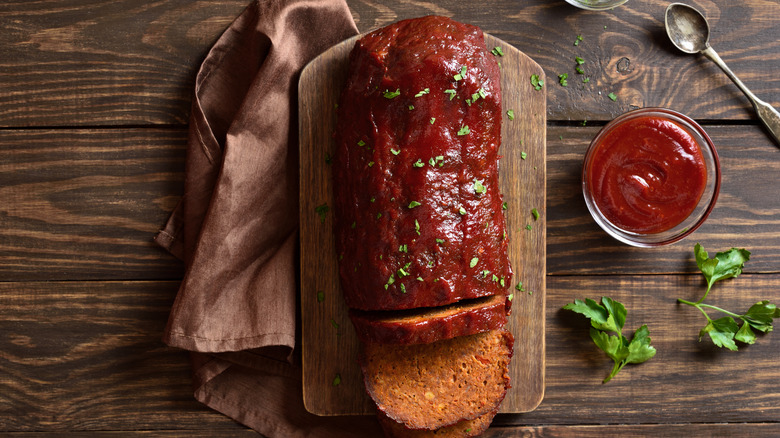 Meatloaf on a wooden board, with herbs, a bowl of sauce, and a cloth on the side
