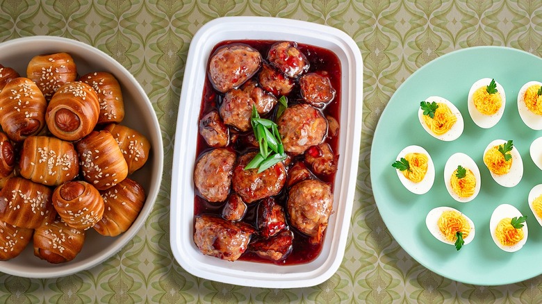 Composite image of pigs in a blanket, grape jelly meatballs, and deviled eggs on a vintage wallpaper background