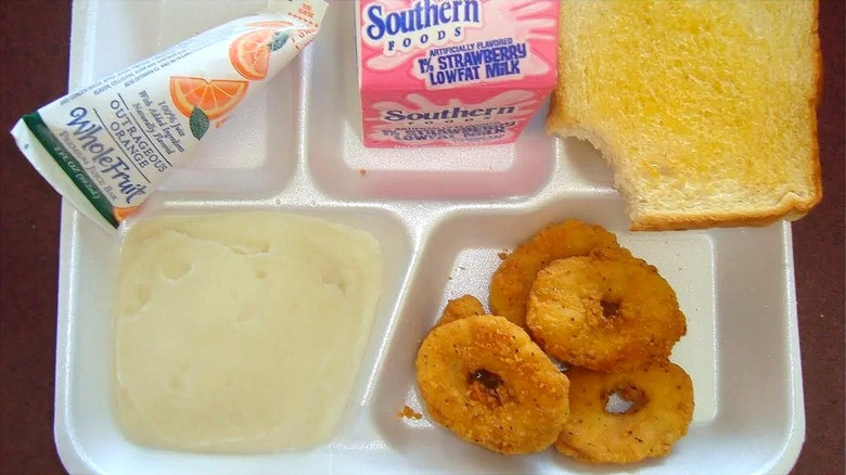 Styrofoam lunch tray with chicken rings, mashed potatoes, buttered bread, strawberry milk, and an orange push pop