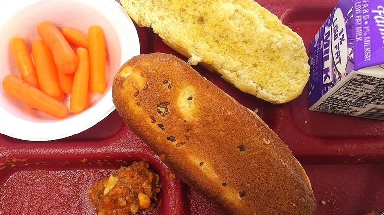 Close-up of a school lunch tray with a buttered hot dog bun, small bowl of baby carrots, and a carton of low-fat milk