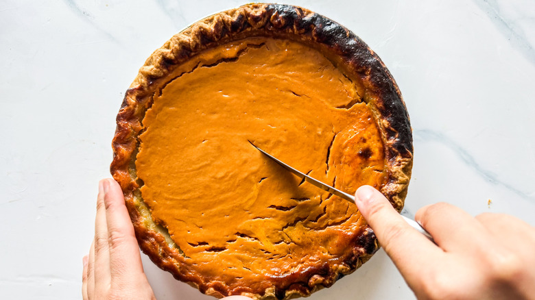 Person slicing into a pumpkin pie placed on marble surface