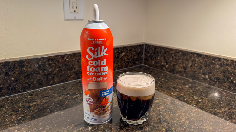 A canister of Silk oatmilk maple brown sugar cold foam next to a glass of coffee with creamer on top