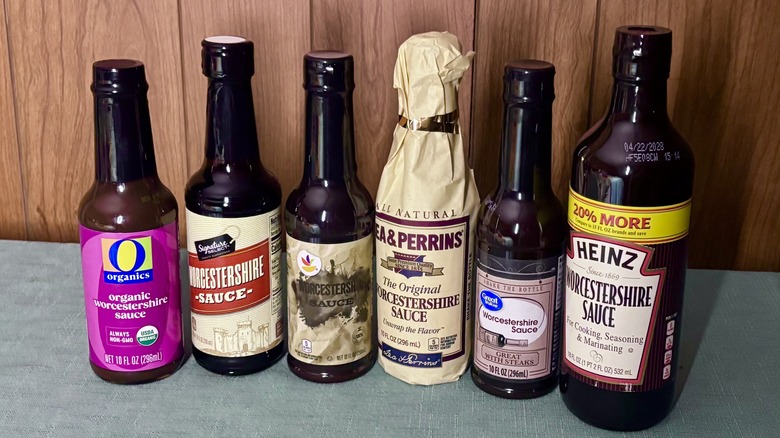 A row of Worcestershire sauces, from left - O Organics, Signature Select, Stop & Shop, a paper wrapped Lea & Perrins, Great Value, and Heinz in a 20% larger bottle than the rest