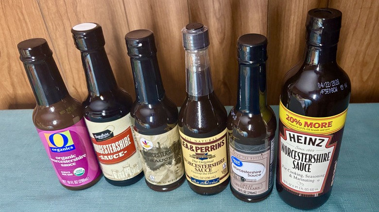 A row of Worcestershire sauces, from left - O Organics, Signature Select, Stop & Shop, an unwrapped bottle of Lea & Perrins, Great Value, and Heinz in a 20% larger bottle than the rest