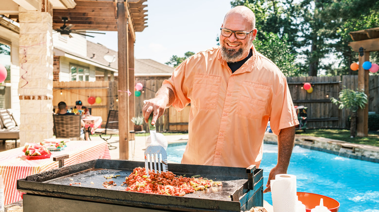 Man frying meat on an outdoor griddle with a pool in the background