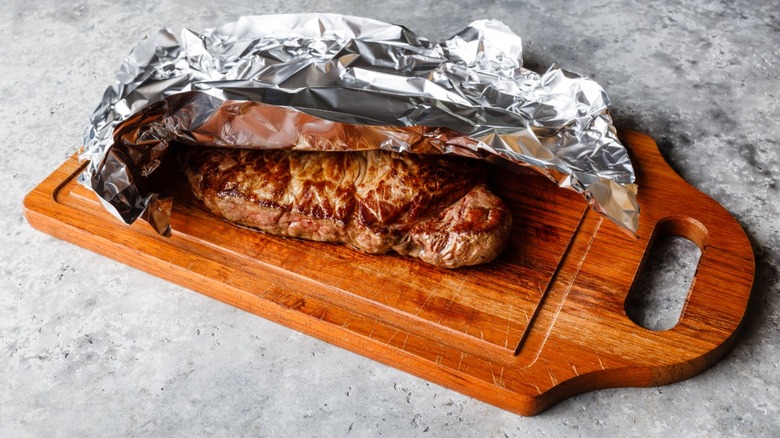A steak resting on a wooden cutting board slightly covered in tin foil