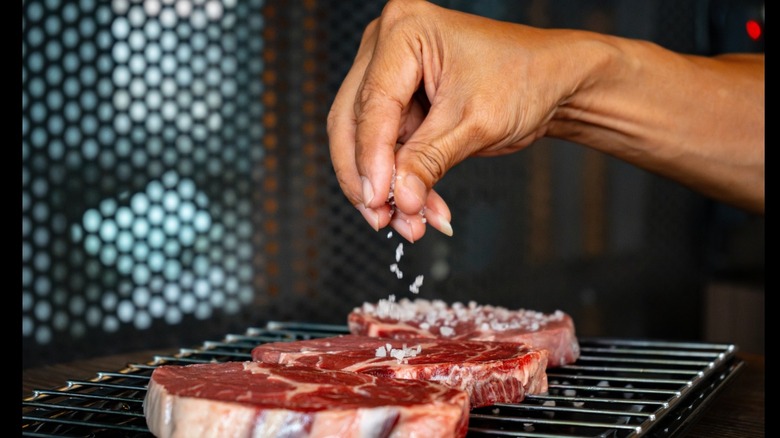 A hand generously seasoning steaks with flaky salt