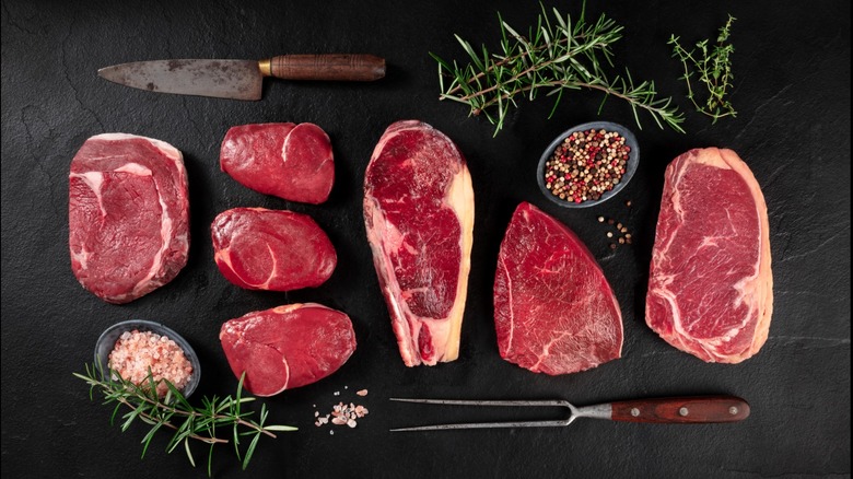 Different cuts of steaks on a black background next to tools and sprigs of rosemary and thyme