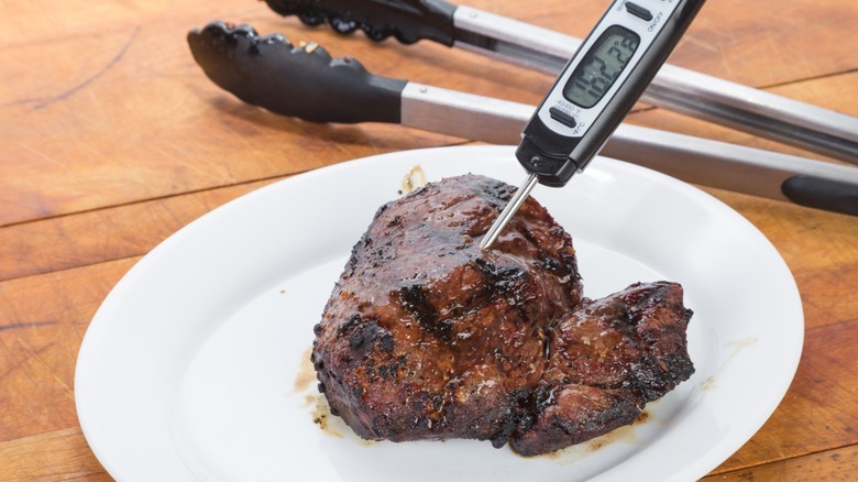 Steak on a white plate with a meat thermometer and tongs in the background