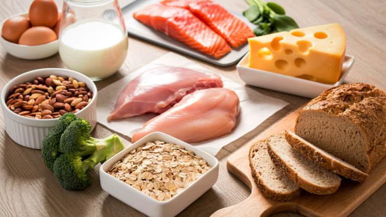 Fresh food, including oatmeal, broccoli, almonds, raw chicken, Swiss cheese block, salmon, bread, eggs, spinach, and a glass of milk, are laid out on a wooden table.