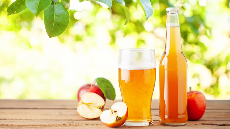 A full glass of hard apple cider sits next to a full clear glass bottle of the same beverage; on a picnic table surrounded by halved and whole apples