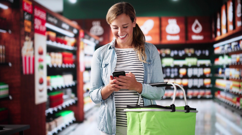 Woman smiling while looking at her smartphone and holding a grocery store basket in the supermarket