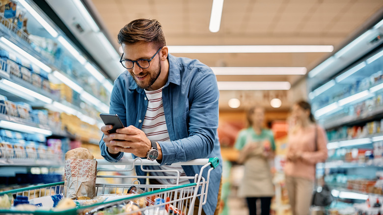 Man using his smartphone while leaning on a full grocery store shopping cart