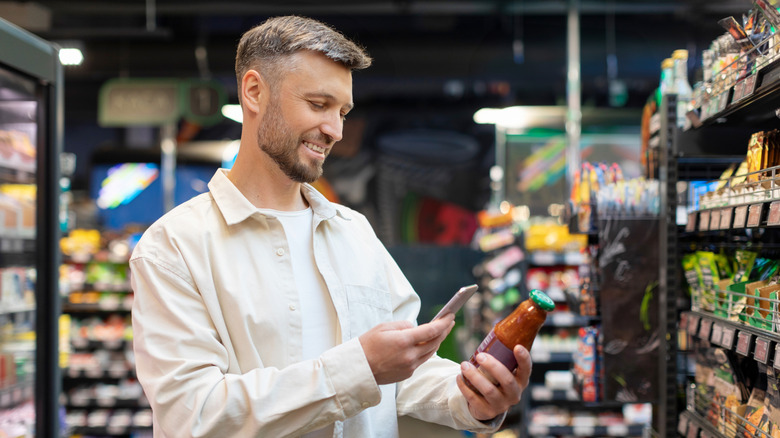 A man holding a jar of salsa and looking at his phone in the supermarket aisle